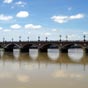 Nous allons franchir le pont de Pierre qui conduit au centre de Bordeaux. L'absence de pont sur la Garonne a longtemps t considre par les Bordelais comme une dfense naturelle face  l'ennemi. Depuis la ralisation du premier ouvrage en 1821, le pont de Pierre, le dsenclavement de la ville a t une priorit vitale. Aprs la passerelle Eiffel en 1860, il faudra nanmoins attendre plus d'un sicle pour construire un nouveau pont. Il relie la rive gauche au quartier de la Bastide. Premier pont sur la Garonne, il fut construit sur ordre de Napolon Ier entre 1810 et 1822, d'aprs les plans de Claude Deschamps. Il mesure 486 m de longueur et 19 m de largeur. Les premires assises de la maonnerie des piles ont t mises en place grce  des caissons compartiments de 23 m de longueur, de 7,40 m de largeur et de 6 m de hauteur. Durant douze ans les btisseurs durent faire face  de nombreux problmes en raison du courant trs fort  cet endroit-l. C'est grce  une cloche  plonge emprunte aux Britanniques que les piliers du pont purent tre stabiliss. Il compte dix-sept arches (nombre de lettres dans le nom de Napolon Bonaparte). Sur les cts, chaque pile de briques est rehausse d'un mdaillon blanc en l'honneur de l'empereur. Il porte aussi les petites armoiries de la ville (les trois croissants entrelacs). Ce pont fut le seul jusqu' la construction du pont Saint-Jean en 1965.